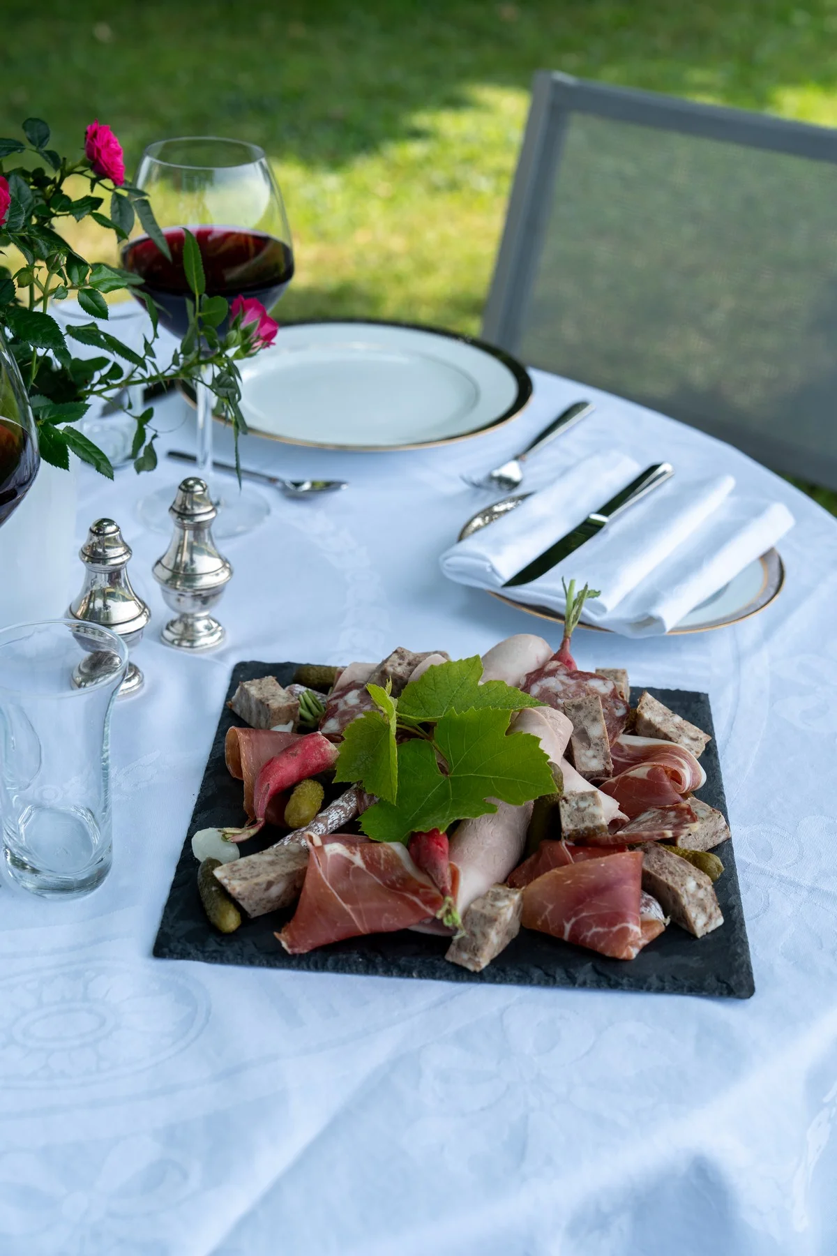 Platte mit handwerklicher Charcuterie und Gläsern mit rotem Beaujolais auf einem blumengeschmückten, im Garten gedeckten Tisch